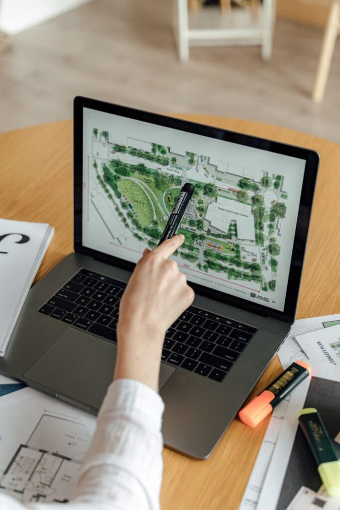 pexels-photo-5244002-5244002 An architect reviews landscape design plans on a laptop, using highlighters and documents at a wooden desk.